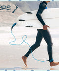 Surfer walking on beach carrying board with Storm 8mm Surfboard Leash 8 Ft Premium Leg Rope attached, Koru Dark Blue