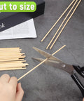 Cutting 150 Piece Smooth Wooden Dowel Rods 1/8 By 12 Inches for DIY crafts with scissors on a gray surface.