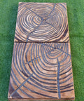 Two rectangular rustic wood planks laying side by side on green grass, each featuring engraved concentric circles and lines.