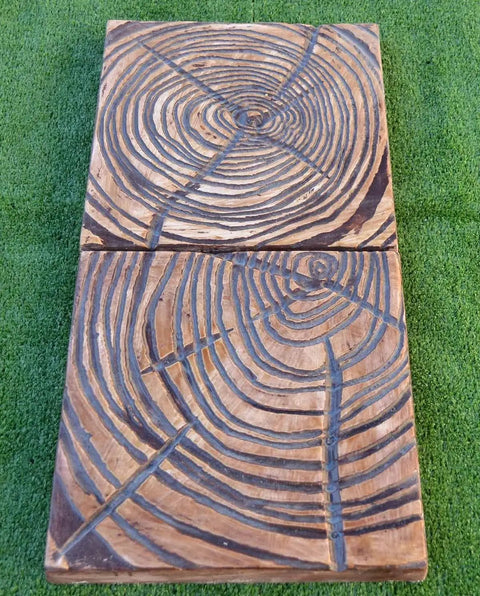 Two rectangular rustic wood planks laying side by side on green grass, each featuring engraved concentric circles and lines.