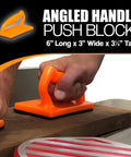 Close-up of orange angled handle push block being used on a wood surface from the Five Piece Woodworking Push Block And Push Stick Set In Safety Orange For Table Saws Router Tables Jointer And Band Saw Keeps Your Fingers Safe While You Push Through Cuts And Tackle Any Workshop Job.