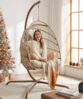 Woman sitting on an Indoor Outdoor Wicker Hanging Egg Chair With Stand Cozy Patio Swing Hammock Egg Shell Chair With Cushions 350 Lb Capacity Sturdy Porch Swing For Backyard Balcony Beige in a living room setting, with a Christmas tree in the background.