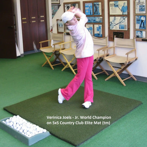 Child practicing golf swing indoor on a 5x5 Country Club Elite Mat, surrounded by chairs and golf balls in a tray.