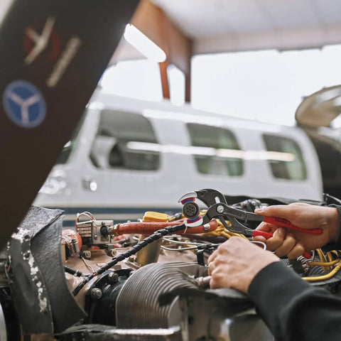 Mechanic using Pipe Gripping Pliers With Replaceable Plastic Jaws For Soft Surfaces on aircraft wiring connector, plane in background.