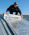 Man applying No Hassle Easy Peel And Stick Asphalt Roofing Roll With Reflective Foil Backing For Heavy Duty Waterproof Leak Patch And Summer Heat Deflector 39 By 16 Feet on a metal roof.
