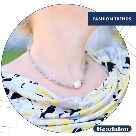 Woman wearing a beaded necklace with a large pearl pendant, against a floral patterned dress in an outdoor setting.
