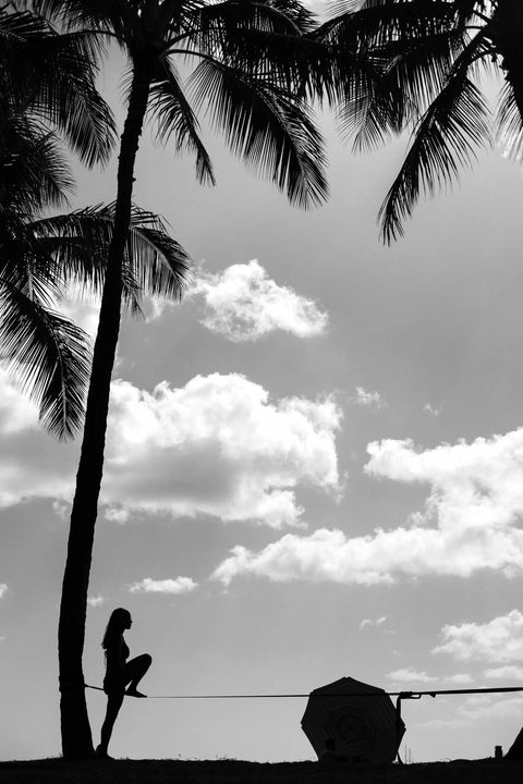 Silhouette of a person balancing on Infinity Slackline Kit With 100 Foot Longline, palm trees in background, cloudy sky.