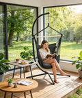 Person relaxing in a Chill Macrame Hammock Chair With Two Cushions For Indoor Outdoor Use Hanging Rope Swing Chair Supports Up To 350 Lbs Anti Slip Spreader Bar And Pocket Black on a patio.