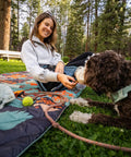 Person sitting on a colorful mat with a dog in a park, enjoying a day outside.
