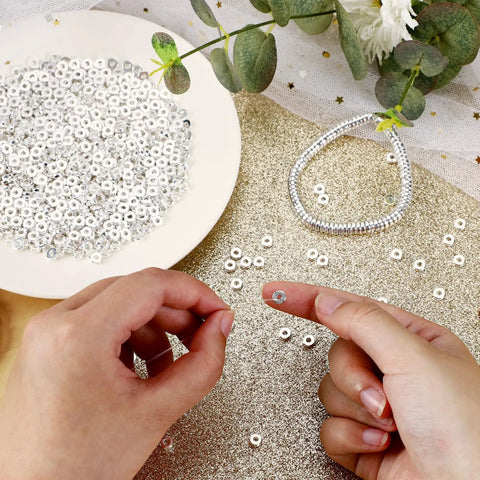 Hands holding six millimeter flat round spacer beads pack of 1500 silver rondelle beads for jewelry making on gold surface with greenery.