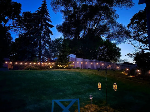 Solar Powered Outdoor String Lights 48 Ft Commercial Grade Waterproof Patio Lights With 15 Shatterproof LED Edison Bulbs glowing in a backyard at night.