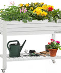 Elevated Garden Bed Kit With Legs And Wheels Large Resin Planter Box Stand With Drain Plug And Storage Shelf Perfect For Plants Vegetables Fruits Outdoor Durable Grayish White filled with flowers and gardening tools on shelf.