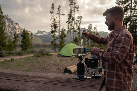 Man using Portable Propane Stove With 10,000 BTU Burner Chrome Anti-Clog Design in a campsite with mountains in the background.