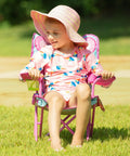 Child sitting in Cutie Pie Butterfly Foldable Kids Chair For Lawn And Camping, pink frame, enjoying outdoors in sun hat.