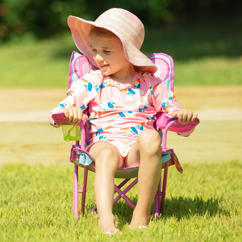 Child sitting in Cutie Pie Butterfly Foldable Kids Chair For Lawn And Camping, pink frame, enjoying outdoors in sun hat.