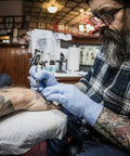 Person with tattoos receiving new tattoo in a studio, tattoo artist in plaid shirt and gloves focused on detail work.