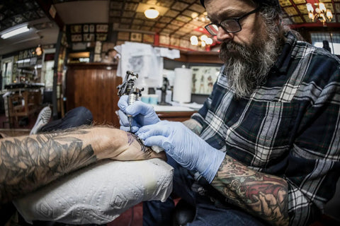 Person with tattoos receiving new tattoo in a studio, tattoo artist in plaid shirt and gloves focused on detail work.