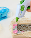 AdTech hot glue gun applying clear 4-inch mini glue sticks to lace on wood; blue tape dispenser in background.
