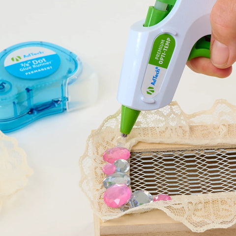 AdTech hot glue gun applying clear 4-inch mini glue sticks to lace on wood; blue tape dispenser in background.