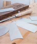 Cabinet Scraper Burnisher With Six Piece Card Scraper Woodworking Set Including Three Rectangle Scrapers One Beveled Curved Concave Gooseneck for smoothing wood surfaces on a workbench.