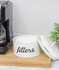 Basket Style White Enamel Coffee Filter Holder on kitchen counter, open with filters inside, next to coffee maker and plant.