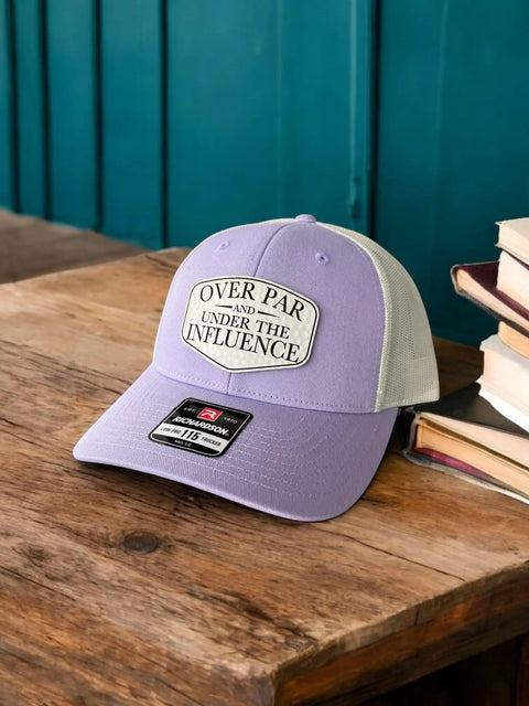 Over Par And Under The Influence Golf Trucker Hat For Women With Dimpled Ball Patch Purple Lilac Birch Color 6 Panel Structured Richardson 115 Cap Funny Golf Accessory displayed on table