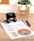 Built Different Two Hole Punch Crushes Twenty Sheets Like Nothing Else With Smart Touch Low Effort Grip And Tight Accuracy Alignment Track For Quick Paper Organizing Jobs In Sleek Black Gray on desk with report.