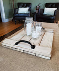 Rustic wooden tray with black handles, mason jar candles, and brown furniture in background.