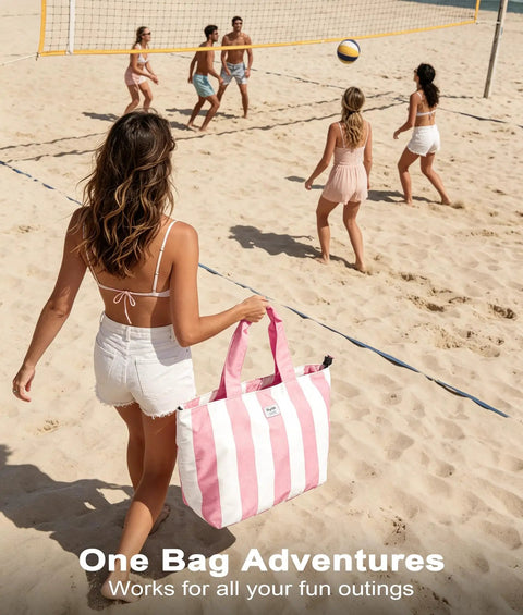 Woman walking on beach holding pink striped bag, volleyball game in background.