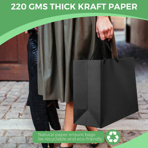 Person carrying an Extra Large Black Gift Bag with handles, sized 16x6x12 inches, showcasing thick kraft paper near cobblestone street.