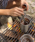 Person using 9.4 inch long spork and spoon set for camping and hiking with deep reach stainless steel utensils on campfire.