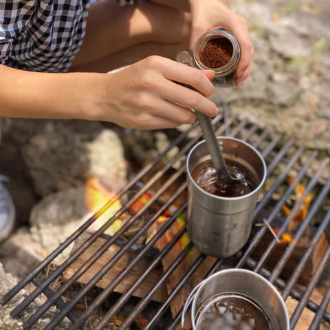 Person using 9.4 inch long spork and spoon set for camping and hiking with deep reach stainless steel utensils on campfire.