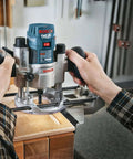 Bosch palm router on wooden workbench, blue body with depth gauge, hands gripping dual handles.