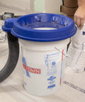 5 Gallon Bucket Dust Collector With High Powered Cyclone Separator And 2 Inch Vacuum Hose Fitting For Serious Jobsite Cleanup with a blue lid and vacuum hose.