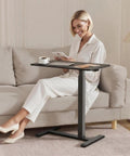 Mobile Standing Desk With Wheels Height Adjustable Pneumatic Laptop Desk Overbed Table in black beside a woman on a sofa.