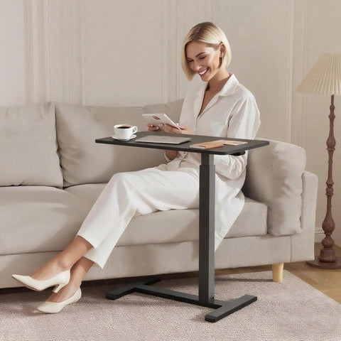 Mobile Standing Desk With Wheels Height Adjustable Pneumatic Laptop Desk Overbed Table in black beside a woman on a sofa.