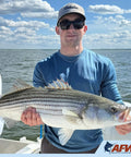 Angler holding a large striped bass on a boat with AFW logo in corner, with lake and sky in background.