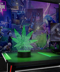 3D Illusion Night Light With Remote, 16 Color Changing LED Lamp in marijuana leaf shape on gaming desk.