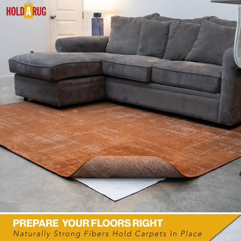 Non Slip Rug Pad 6 Foot By 9 Foot One Eighth Inch Thick For Hardwood Vinyl Tile And Laminate Floors Keeps Area Rugs Secure Safe Indoors With Extra Grippy Comfort, visible under corner of brown area rug in living room with grey sofa.