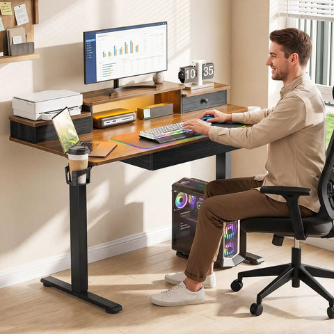 A man seated at an Electric Standing Computer Desk With RGB Lighting And Drawers For Home Office Gaming Setup, working on a laptop.