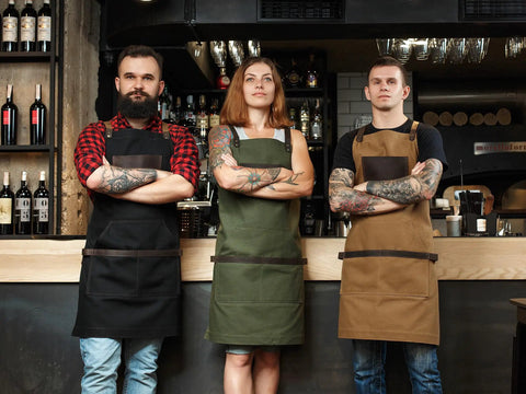 Three individuals in black, green, and brown aprons stand confidently behind a bar, showcasing Black Canvas And Leather Apron For Chefs Bartenders Baristas Barbers And Grill Masters Handmade In USA Durable Kitchen Workwear With Pockets Ideal For Pro Chefs Home Cooks.