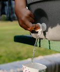 Close-up of a hand using the spigot on a Five Gallon Portable Sports Cooler And Water Dispenser in Gray and Black outdoors.