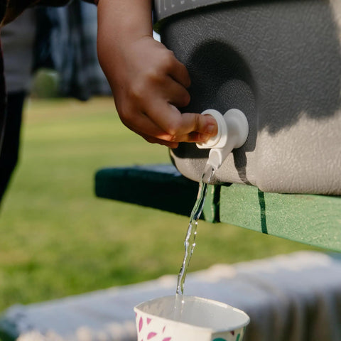 Close-up of a hand using the spigot on a Five Gallon Portable Sports Cooler And Water Dispenser in Gray and Black outdoors.