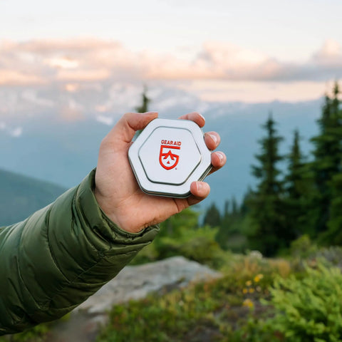 Hand holding the All Weather Tent And Gear Repair Kit With Seam Grip WP 0.25 oz, in metal tin, shown outdoors.