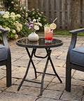 Compact Outdoor Folding Side Table With Tempered Glass Top And Heavy Duty Steel Frame For Backyard Hangouts, holding drinks and flowers.