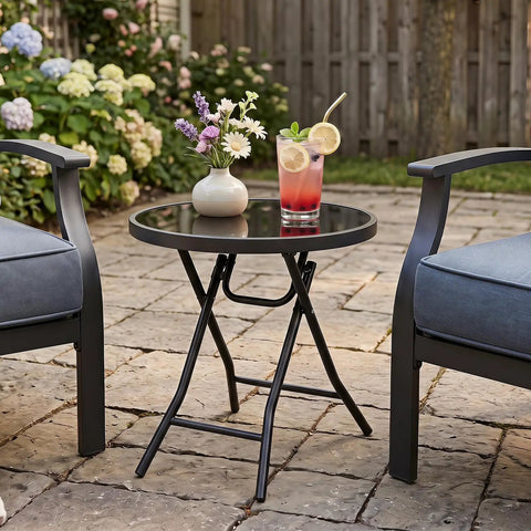 Compact Outdoor Folding Side Table With Tempered Glass Top And Heavy Duty Steel Frame For Backyard Hangouts, holding drinks and flowers.