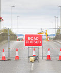 Road closed scene with orange traffic cones and a sign, barrier in place, leading to a blocked-off street under cloudy skies.