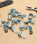 Heavy Duty One Hole 1/2 Emt Straps 100 Pack Zinc Plated Steel Conduit Clamps for Electrical Tubing scattered on wood surface.