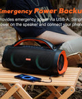 Person sitting outside with Extreme Boom Pro 100W Outdoor Speaker, orange accents, on a wooden table, phone connected via USB.