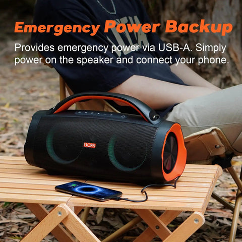Person sitting outside with Extreme Boom Pro 100W Outdoor Speaker, orange accents, on a wooden table, phone connected via USB.
